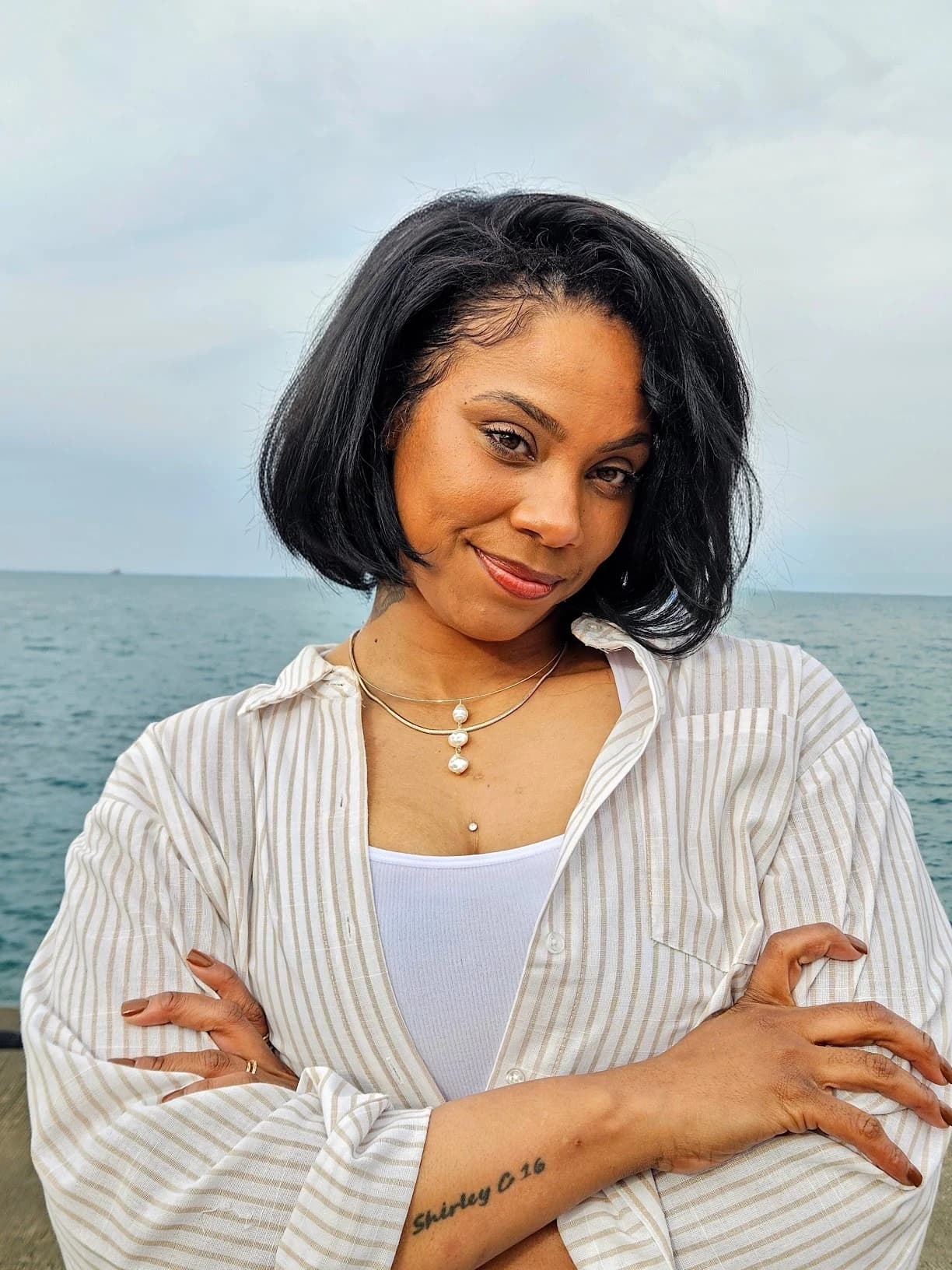 Kalisha at the lakefront in a striped shirt, with her arms folded